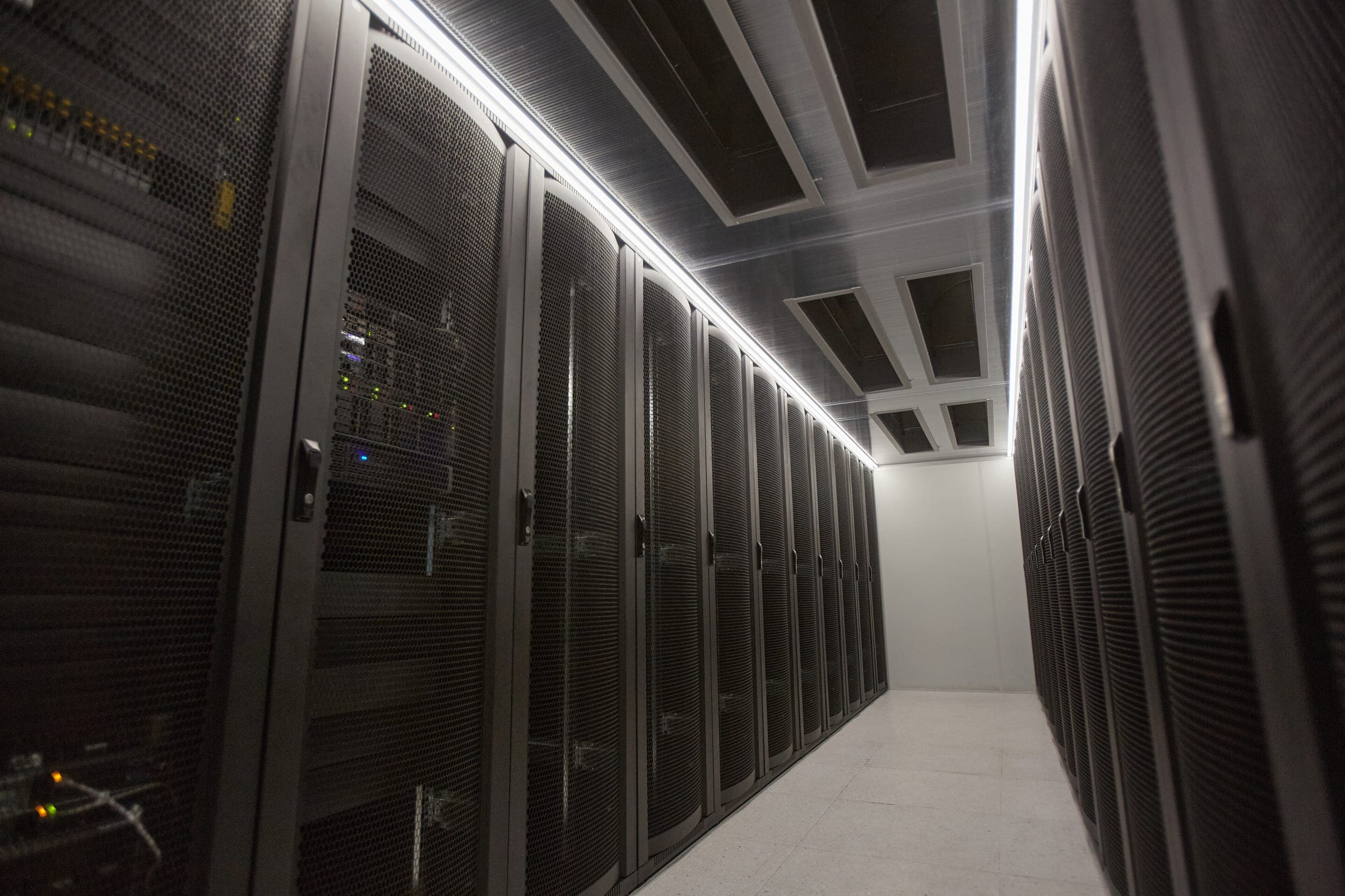 An aisle of server racks in a data center, lit from the ceiling
